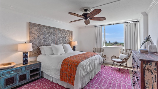 A bedroom with a bed, two bedside tables with lamps, wicker chairs, a dresser, and a window with curtains, all decorated in a mix of rustic and modern styles.