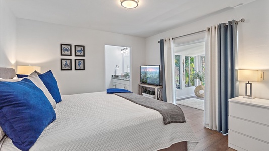 A neatly arranged bedroom with a bed, blue and white bedding, bedside tables with lamps, wall art, a TV, and a view into an adjacent room with large windows.