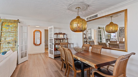 A dining room with a wooden table and six chairs, a large mirror on the wall, and two hanging pendant lights. The room features wooden flooring and double doors leading to an adjacent room.