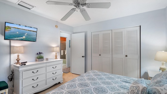 A bedroom with a patterned bedspread, dresser, wall-mounted TV, ceiling fan, and closet. The room has two lamps on the dresser and an open door leading to a bathroom.