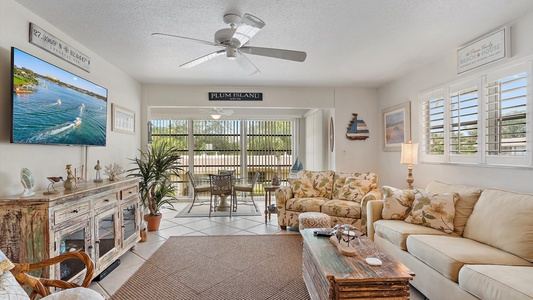 A living room featuring beige furniture, a rustic wooden coffee table, a large TV displaying a water scene, and a sign reading "Plum Island." Large windows reveal an outdoor area with chairs and a table.