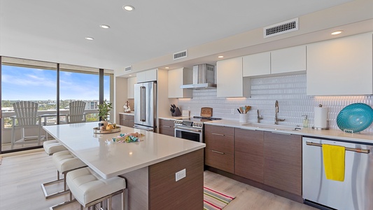 Modern kitchen with white and wooden cabinets, a large island with barstools, stainless steel appliances, and a view of a balcony with outdoor seating.