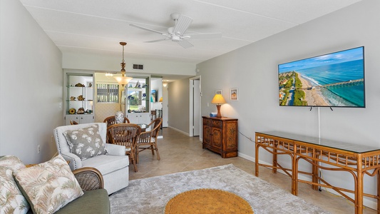 A cozy living room with a white armchair, patterned cushions, a rug, wall-mounted TV showing a beach, and a dining area with a chandelier in the background. The room has light-colored walls.