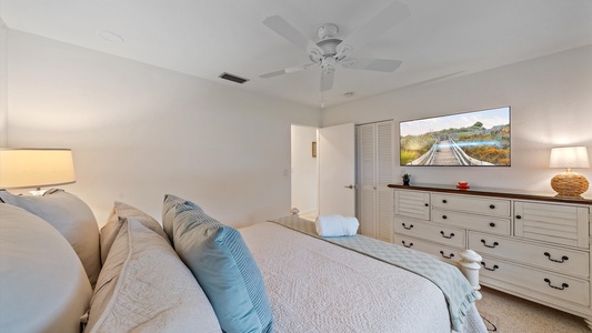 A neatly arranged bedroom with a large bed, white bedding, and blue accent pillows. A ceiling fan is above, a dresser with a TV and lamp on top is against the wall, and a doorway is visible.