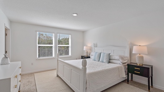 A bright bedroom with a white bed, two black nightstands with lamps, a white dresser with vases, and a window with blinds allowing natural light to enter the room.