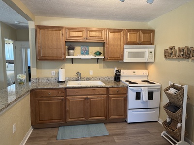 A compact kitchen with wooden cabinets, a granite countertop, a white electric stove and microwave, a white sink, and a paper towel holder. There are decorative items on shelves and a mat on the floor.