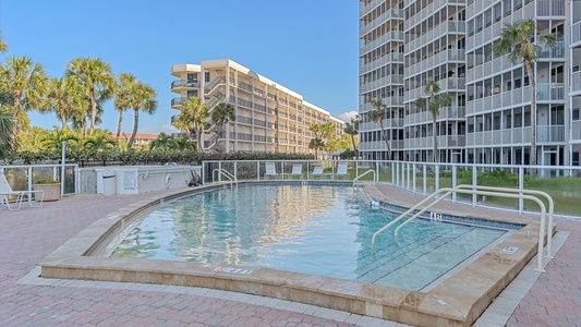 Outdoor swimming pool surrounded by a paved deck area, with several multi-story buildings and palm trees in the background. Safety railings are positioned around the pool for easy access.
