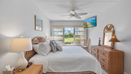 A bedroom featuring a bed with white linens, two nightstands with lamps, a dresser with a mirror and lamp, a wall-mounted TV, and a window with a view of a green lawn and sky.