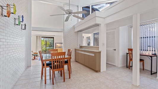 Spacious white brick dining area with wooden table and chairs, ceiling fan, large windows, and adjacent open living room featuring sliding doors to an outdoor area.