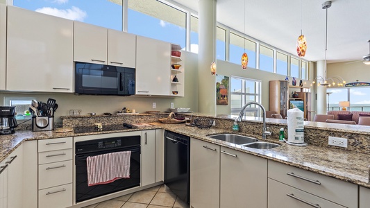 Modern kitchen with granite countertops, white cabinets, a double sink, and built-in appliances. Sunlit with large windows and pendant lights. Dining and living areas visible in the background.