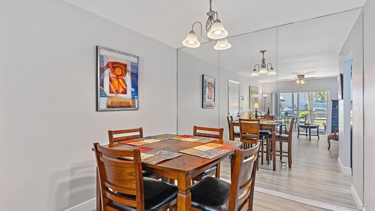 A dining room with a wooden table and four chairs, a mirror on one wall, abstract artwork, and a view into a living area with additional seating and a window.