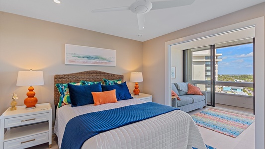 A modern bedroom with a bed featuring white and blue bedding, flanked by two lamps. A sitting area with a couch and a colorful rug is visible through an opening. A balcony with a view is in the background.