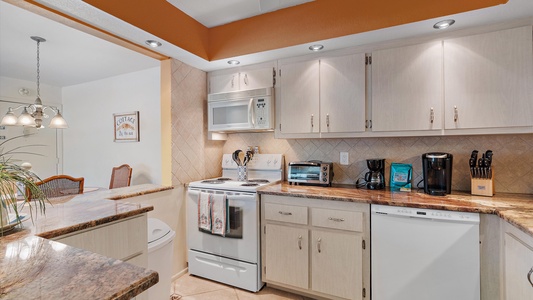 A modern kitchen with white cabinets, brown granite countertops, a white stove, microwave, toaster, coffee maker, dishwasher, and a dining area visible in the background.