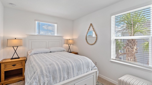 A small bedroom with a white bed, two wooden nightstands with lamps, a woven round mirror on the wall, and windows with blinds showing a view of a palm tree outside.