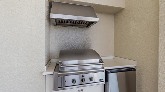 A stainless steel outdoor grill with a closed lid is positioned under a vent hood, beside a small refrigerator, all set against a textured beige wall.