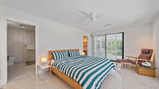 A modern bedroom with a striped bedspread, a bedside table with a lamp, a chair, a ceiling fan, and sliding glass doors. The room also features an attached bathroom with tiled walls.