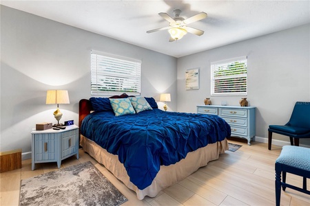 A bedroom with a navy blue bedspread, two nightstands with lamps, a ceiling fan, light-colored walls, two windows with blinds, a blue dresser, a blue chair, and light wood flooring.