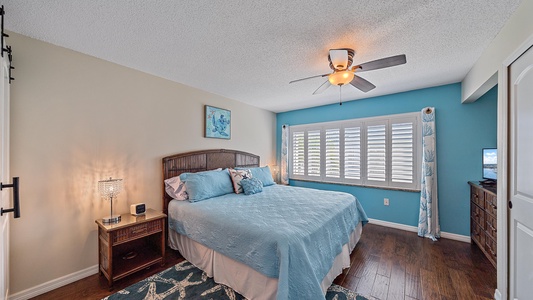 A bedroom features a bed with light blue bedding, two bedside tables with lamps, a ceiling fan, a dresser, a rug, and a window with plantation shutters. The walls are beige and blue.