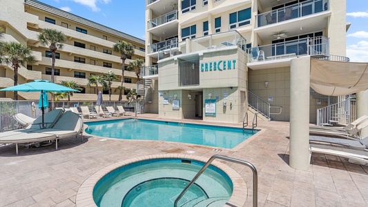 Outdoor pool area beside a multi-story building with lounge chairs, umbrellas, and a hot tub. The building has balconies with glass railings and a sign that reads "Crescent.
