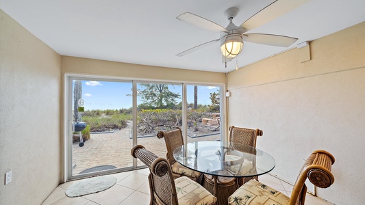 A small dining area with a round glass table and four wicker chairs, situated next to large sliding glass doors that open to a patio and garden view. A ceiling fan is mounted above the table.