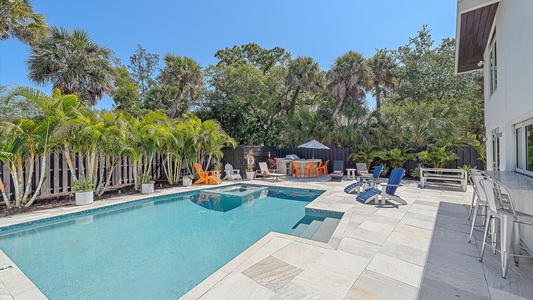 Outdoor pool area with a blue rectangular pool, spa, lounge chairs, palm trees, outdoor seating, and dining areas with umbrellas, all under a clear blue sky.