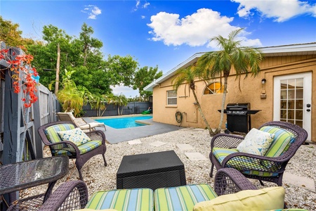 Outdoor patio area with striped cushioned chairs, a grill, and a swimming pool beside a tan house. The sky is partly cloudy with trees in the background.