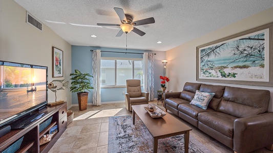 A living room with a ceiling fan, leather couch, armchair, coffee table, flat-screen TV, wall art, and a large window with curtains. The room includes plants and a patterned rug on tiled flooring.