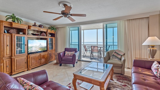 A living room with wooden furniture, a TV, two armchairs, a glass coffee table, a ceiling fan, and a balcony with a sea view.