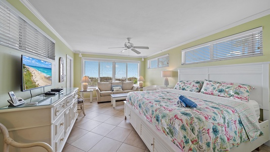 A brightly lit bedroom with a floral patterned bed, a TV, a small seating area, and a view of a beach through large windows. The room features light green walls, tiled floors, and white furniture.