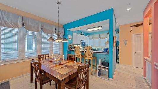 A dining area with a wooden table and four chairs beside a kitchen with a blue wall. The kitchen has two bar stools at the counter. There are large windows with blinds in the background.