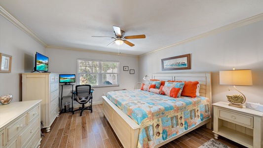 A bedroom with a bed covered in a colorful quilt, two bedside tables, a ceiling fan, a dresser with a TV, and a desk with a chair and computer setup near a window with blinds.