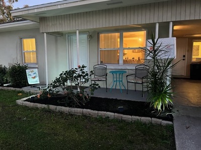 Front view of a single-story house with a small porch that has two chairs and a round table. The porch is decorated with plants and dimly lit from inside. A butterfly sign is visible on the lawn.