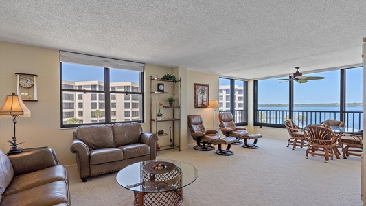 A spacious living room with leather seating, a glass coffee table, and a shelf. Large windows offer a view of a waterfront and surrounding buildings. A dining area with a round table is visible.