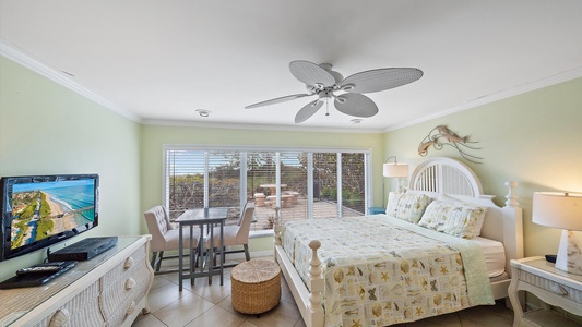 A bedroom with a coastal theme featuring a bed, a small table, a TV on a dresser, a ceiling fan, and a sliding glass door opening to a patio with outdoor furniture.