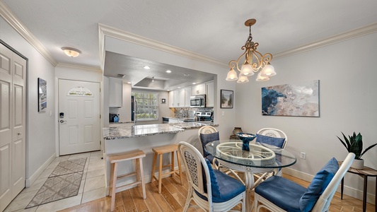 Small kitchen and dining area with a glass table and four chairs, a chandelier, a breakfast bar with stools, and modern appliances. Walls are painted light grey with two framed pictures.