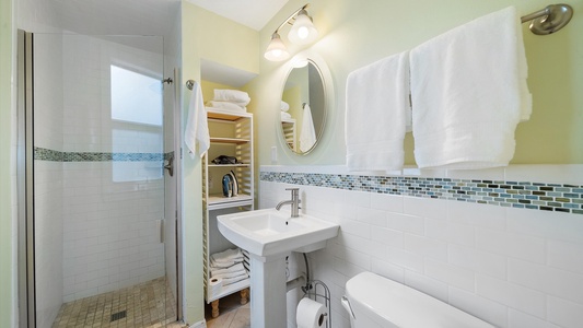 Small bathroom with a shower enclosed by glass doors, pedestal sink, round mirror, toilet, and open shelving unit stocked with towels. Walls have white tiles with a mosaic trim, under soft lighting.