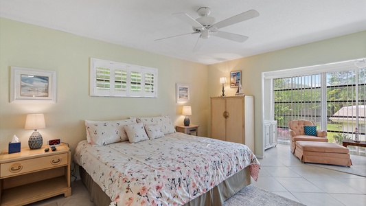 A bedroom with a double bed, floral-patterned bedding, nightstands with lamps, an armoire, a chaise lounge, and a ceiling fan. The room has a window with blinds and appears bright and airy.