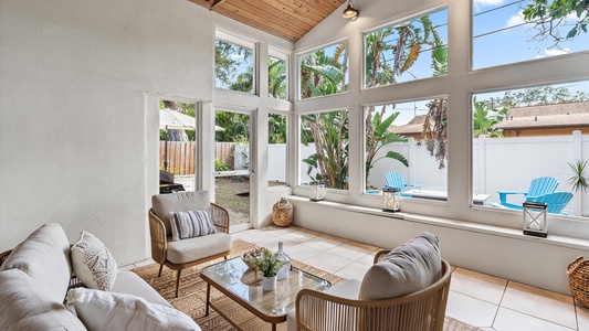 A cozy sunroom with large windows has a seating area including a white sofa, wicker chairs, and a glass coffee table. The room overlooks a backyard with greenery and a pool.