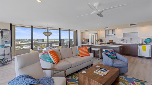 Modern open-concept living room and kitchen with large windows, light wood flooring, a ceiling fan, colorful cushions, and a mix of white and dark wood cabinetry.