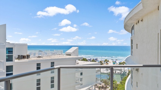 A view of the ocean from a high balcony, showing white buildings, beachgoers, and a bright blue sky with scattered clouds.