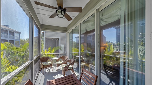 A small screened-in balcony with a ceiling fan, three wicker chairs with cushions, a wooden table, and surrounding lush greenery outside. Large glass doors provide access to the interior.