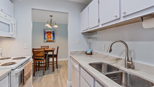 A modern kitchen with white cabinets, a stainless steel sink, and a white stove leads to a dining area with a table and four chairs under a light fixture. A colorful painting hangs on the wall.