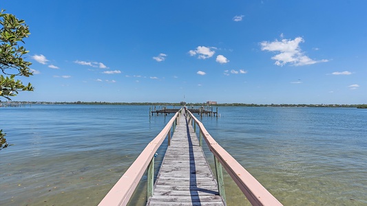 A long wooden pier extends over clear blue water toward a dock, with a few trees to the left and a clear blue sky with some clouds above.