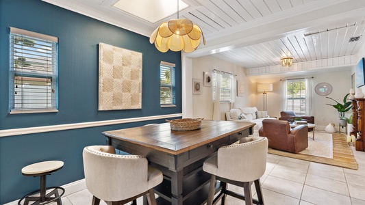 A modern open-concept living and dining area with a wooden dining table, cream-colored chairs, blue accent wall, wicker decor, white paneled ceiling, and a cozy living space with sofas and a brown armchair.