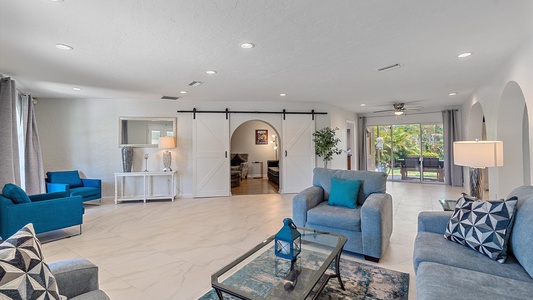 Spacious living room with gray sofas, blue armchairs, a glass coffee table, and a sliding barn door. Large windows and sliding glass doors provide natural light. Modern decor and neutral color palette.