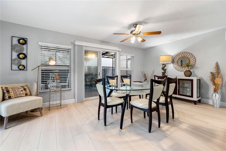 A dining room features a glass table with six chairs, a ceiling fan, a cabinet with decor, a floor lamp, a small cushioned bench, and large windows with blinds and sliding glass door.