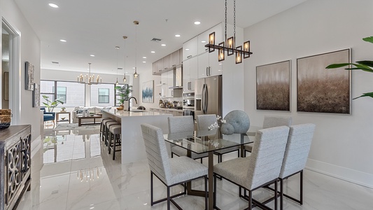 Modern open-concept kitchen and dining area with light-colored walls and floor, glass dining table with cushioned chairs, bar stools at the kitchen island, and contemporary light fixtures.