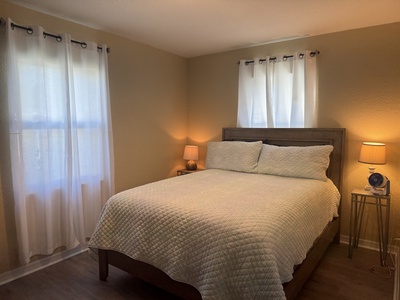 A neatly made bed with white quilts and pillows is positioned against the wall. Two bedside tables with lamps on each side of the bed. Light curtains hang over the windows on adjacent walls.