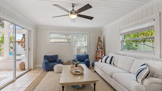 A bright living room features a white couch with striped pillows, two blue armchairs, a beige rug, a wooden coffee table, a ceiling fan, and large windows offering natural light and garden views.