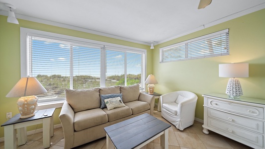 A small living room with green walls, a beige sofa, a white armchair, two lamps on side tables, a white dresser, and large windows with blinds overlooking an outdoor area.